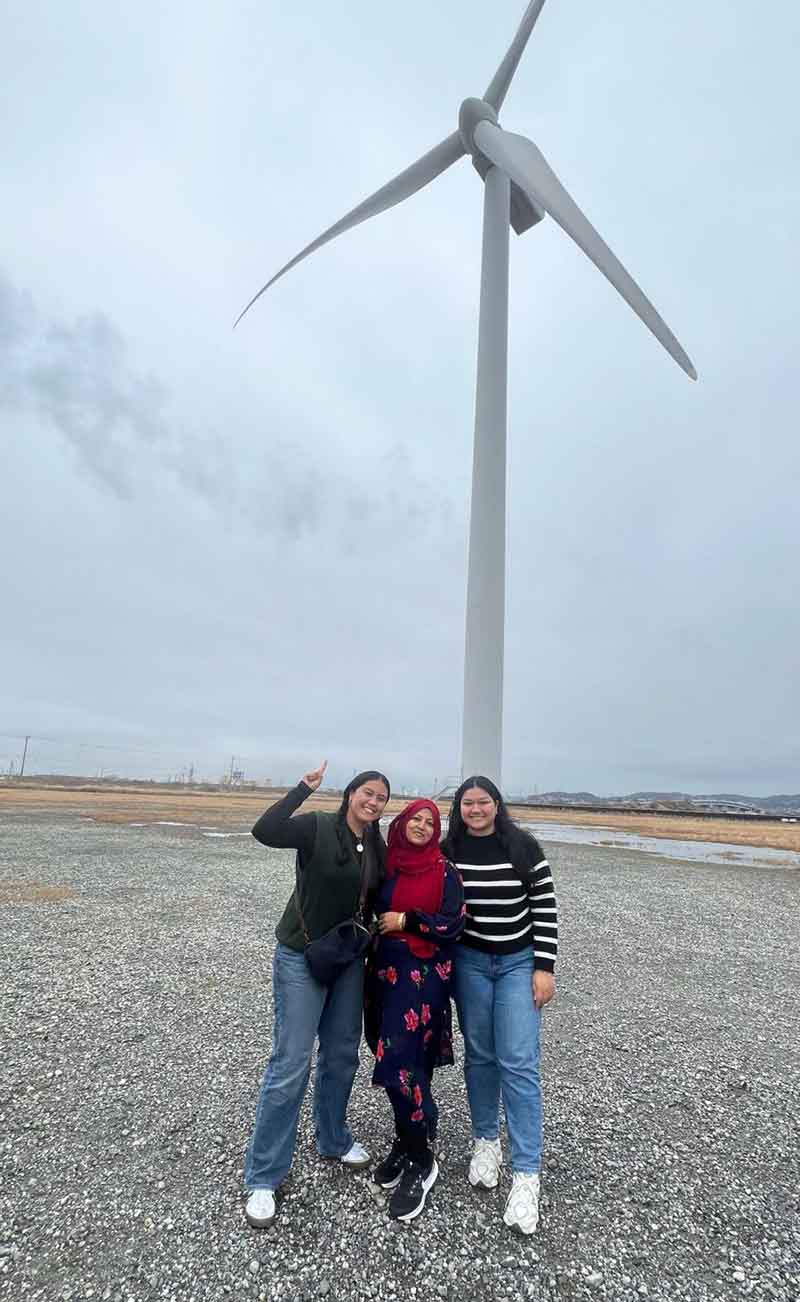 Photo of three women standing in front of a wind turbine in Japan