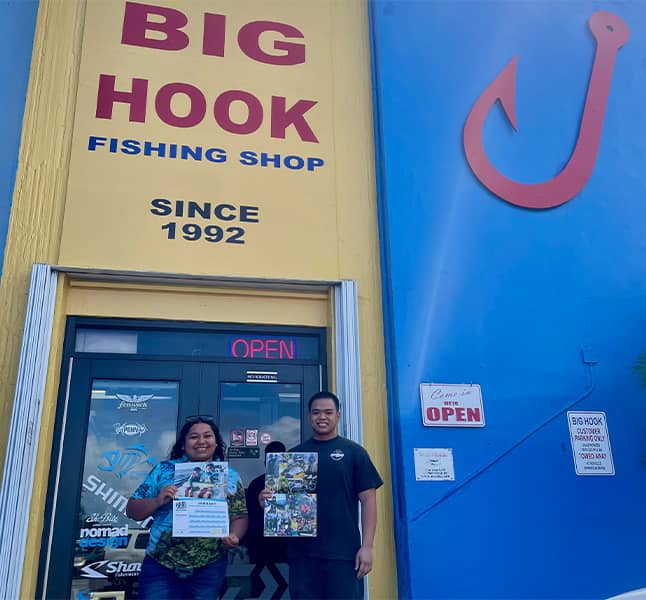 Gabriella Topasna poses with employee in front of Big Hook holding the 2024 Guam Tide Chart Calendar