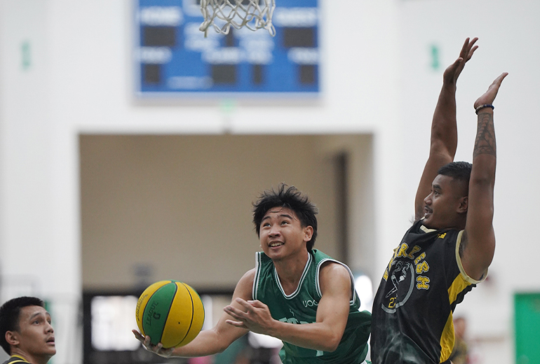 Triton player in green jersey holds ball out under hoop, opponent blocks
