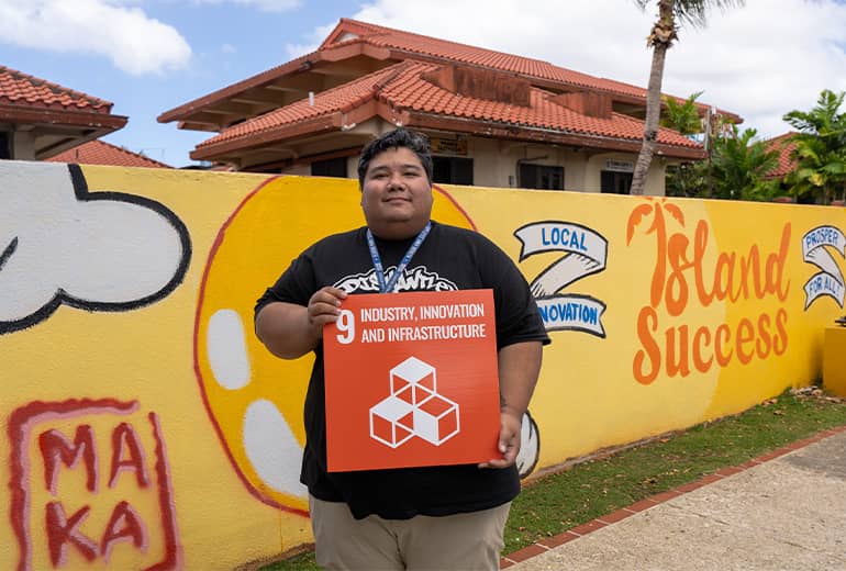 Man stands with G3 poster showing SDG #9 in front of his mural at CHamoru village