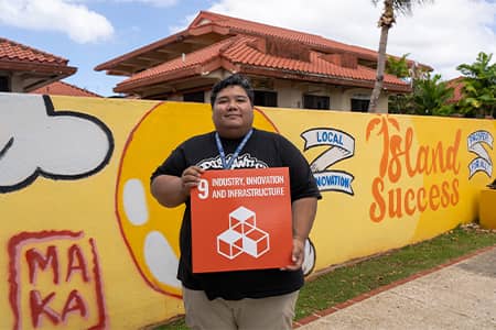 Man stands with G3 poster showing SDG #9 in front of his mural at CHamoru village