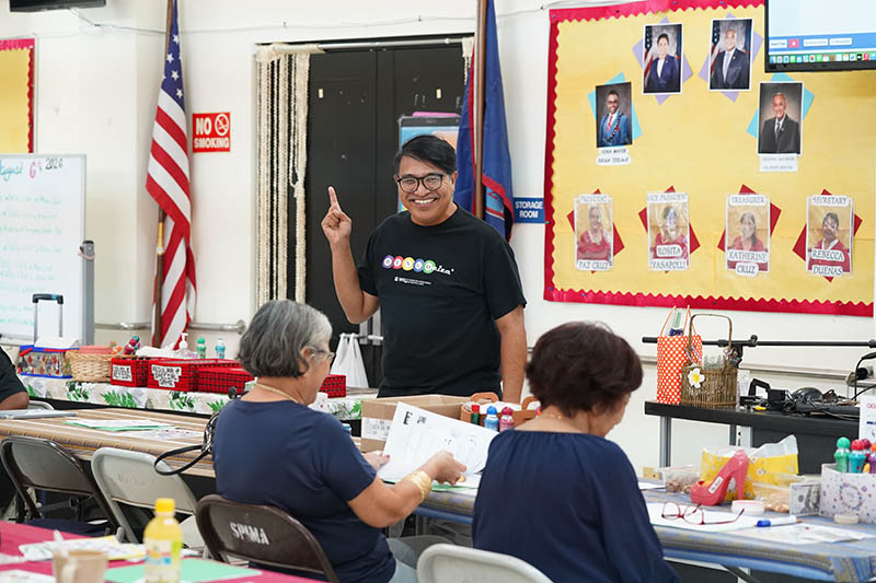 A smiling man standing in front of a room of older adults in a senior center